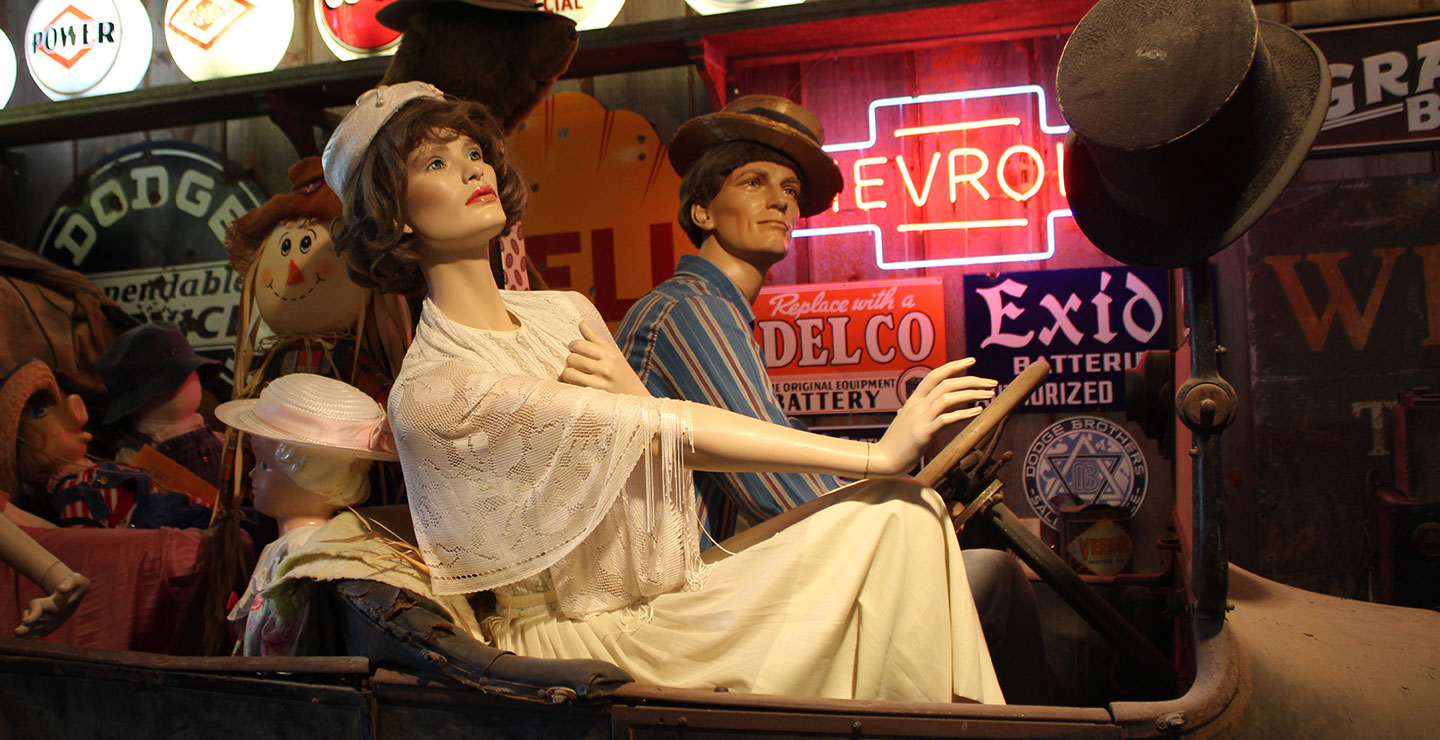 Mannequins in an old car; neon signs are lit behind them