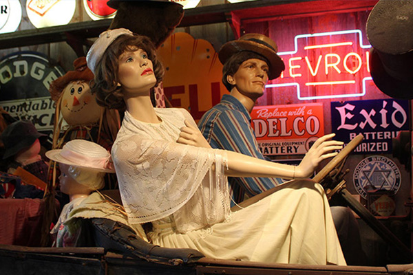 Mannequins in an old car; neon signs are lit behind them